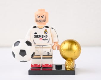 Miniatura do Zinedine Zidane com o uniforme número 5 do Real Madrid e a taça da Copa do Mundo. Uma peça icônica para exibir e presentear seu filho fã de futebol na formatura.