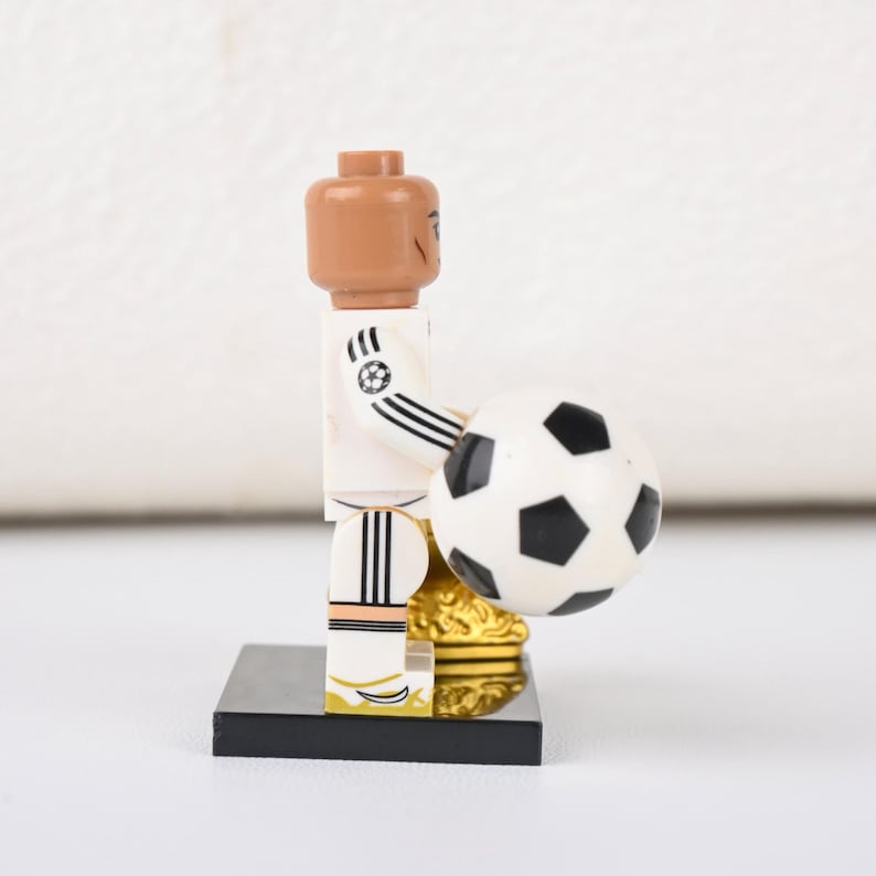 May include: A Lego figurine of a footballer in a white kit with black stripes, holding a black and white football. The figurine has a brown head and is standing on a black base with a gold trophy.