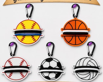 May include: Five circular Game-Day Hair Tie Holders with black carabiners. Designs include a yellow softball, orange basketball, white baseball with red stitching, black and white football, and white volleyball, each holding elastic hair ties.