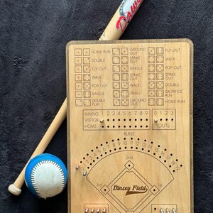 May include: A wooden baseball scorekeeping board with engraved game outcomes, inning trackers, and a field diagram labelled Dinzey Field, accompanied by a light-coloured wooden baseball bat with red Dawgs text and a blue and white baseball.