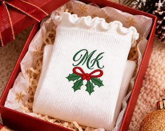 Puede incluir: Una bolsa de tela blanca acanalada con un borde superior con volantes, que presenta un diseño bordado de las letras MK en letra verde sobre un lazo rojo y hojas de acebo verdes, presentada dentro de una caja de regalo roja.