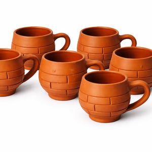 Könnte beinhalten: Ein Set aus fünf terrakottafarbenen Tassen mit abgerundeter Form und einer strukturierten Außenfläche, die an gestapelte Ziegel erinnert. Jede Tasse verfügt über einen geschwungenen Henkel an der Seite und eine weite Öffnung in Erdtönen.