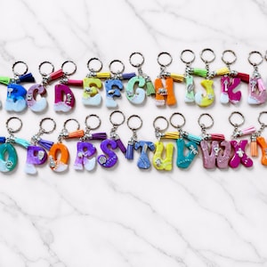 May include: A collection of twenty-six alphabet letter keychains made from colorful resin. Each letter features a unique blend of colors, a small silver-toned charm, and a matching fabric tassel attached to a circular metal split ring keychain.