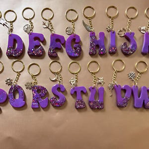 May include: A collection of twenty-six purple resin alphabet keychains arranged in two rows. Each letter features a gradient effect with embedded glitter at the base and is attached to a gold-toned metal keyring with a small decorative charm.