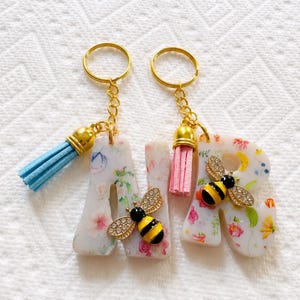 May include: Two resin letter keychains, A and R, featuring a white base with colorful floral patterns. Each letter is adorned with a bee charm with crystal wings and a gold-tone metal keyring. The A has a light blue tassel, and the R has a pink tassel.