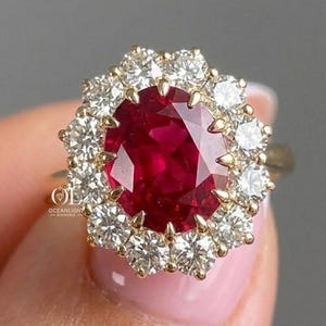 May include: A gold ring featuring a large oval-cut deep red ruby center stone surrounded by a halo of twelve round brilliant-cut white diamonds. The setting is prong-set in yellow gold, and the text OCEANLIGHT DIAMOND is visible on the side.