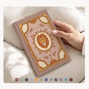 May include: A dusty rose journal with a gold border and a lion illustration. The cover reads "Maria's Journal" and features a lion illustration in a gold oval. The journal is held by a hand.
