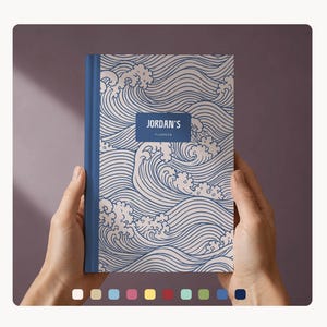 Peut inclure: Un agenda à couverture rigide avec un motif de vagues bleues et blanches. Le dos est bleu uni. La couverture porte le texte "JORDAN'S PLANNER" dans un rectangle bleu marine.