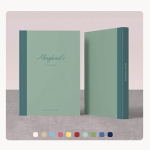 May include: Two sage green planners with dark teal spines. The front planner has the text "Margharet's Planner" in cursive script. The bottom of the planner reads "One Day at a Time". The spine of the second planner reads "Make it Happen".