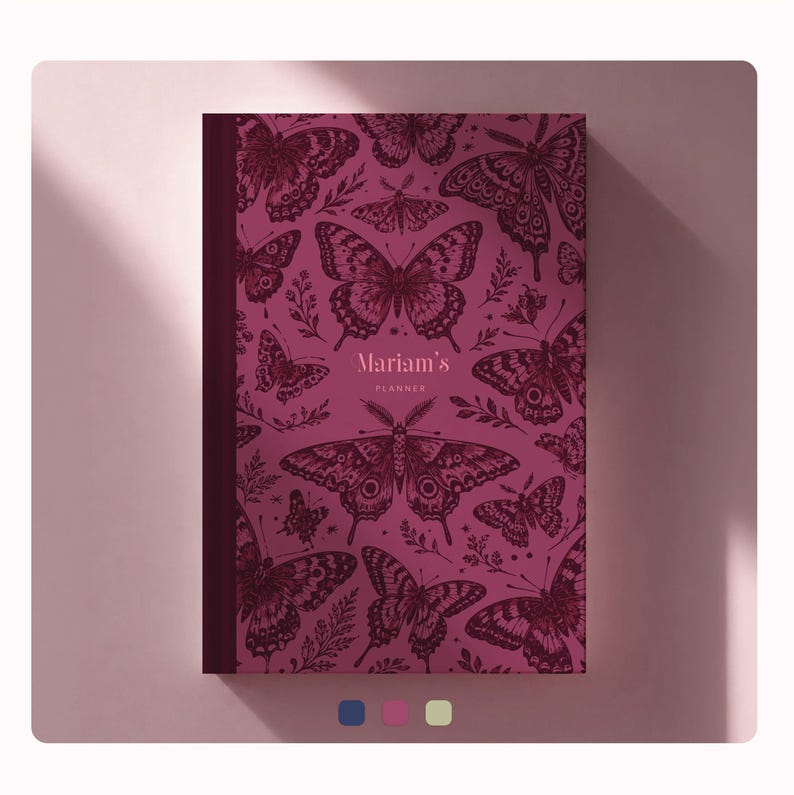 May include: A dark magenta planner featuring a repeating pattern of black ink-style butterflies and foliage. The center of the cover displays the text Mariam's PLANNER in a light pink font. The planner has a solid dark spine on the left side.