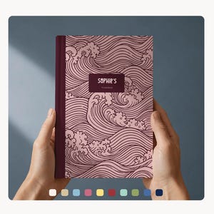 Könnte beinhalten: Ein Planer mit weinrotem Rücken und einem rosa Einband mit einem Wellenmuster. Der Einband trägt den Text "Sophie's Planner" in einem weinroten Rechteck. Der Planer wird von zwei Händen gehalten.