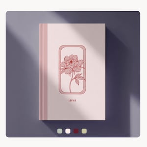 Puede incluir: Un cuaderno rosa claro con una ilustración floral en un marco rectangular. La ilustración es un dibujo detallado de una flor de peonía. El nombre "SOPHIE" está impreso debajo de la flor. El cuaderno tiene una lomo rosa y está sobre un fondo morado.