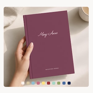 May include: A burgundy journal with the name "Mary-Anne" in cursive script. The words "REFLECTION JOURNAL" are printed at the bottom. The journal is held by a person's hand. A palette of color swatches is at the bottom.