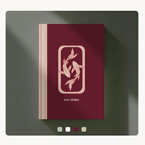 Può includere: Un diario bordeaux con dorso color crema. La copertina presenta un design rettangolare con due pesci koi e la scritta "RILEY BROOKS". Il diario è su una superficie verde.