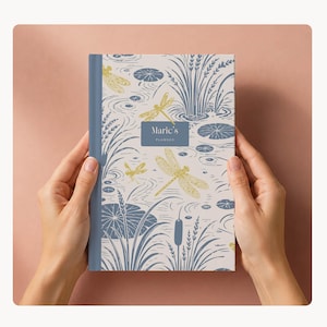 May include: A planner with a dragonfly and lily pad design. The cover features a blue spine and a cream-coloured background with illustrations of dragonflies, lily pads, and reeds in yellow and blue. The text "Maric's Planner" is on the cover.
