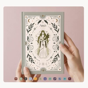 May include: A sage green journal with the text "Anna's Journal" and an illustration of a woman holding wheat. The cover features a decorative border with stars, moons, and floral elements. The journal is held by two hands.