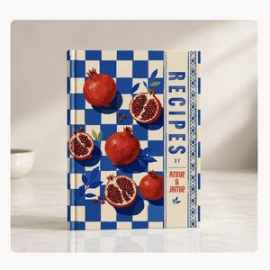 Puede incluir: Un libro de cocina titulado "RECIPES" de Annie & Jamie. La portada presenta un patrón de cuadros azules y blancos con ilustraciones de granadas. El lomo es de color crema con diseños florales azules y el título.