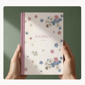 May include: A planner with a floral design and the text "Kathrine's Planner" on the cover. The cover has a light background with pink, blue, and white flower illustrations. The spine is a solid pink colour.