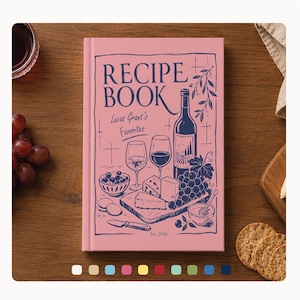 May include: A pink recipe book with the title "RECIPE BOOK" in navy blue, featuring illustrations of wine, cheese, and grapes. The book is titled "Lucas Grant's Favorites" and includes the year "Est. 2026". The book is on a wooden surface.