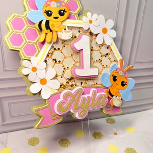 May include: A layered cake topper featuring a honeycomb design with pink and gold glitter accents, two cartoon bees, white daisies, and the number 1. A pink banner at the bottom displays the name Ayla in gold glitter script lettering.