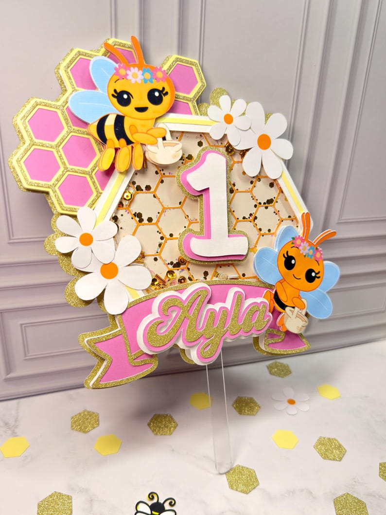 May include: A layered cake topper featuring a honeycomb design with pink and gold glitter accents, two cartoon bees, white daisies, and the number 1. A pink banner at the bottom displays the name Ayla in gold glitter script lettering.