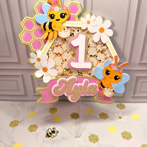 May include: A decorative cake topper featuring a hexagonal shaker filled with gold glitter, a large white number 1, and the name Ayla in pink script. The design includes two cartoon bees, pink honeycomb patterns, and white daisy flowers.