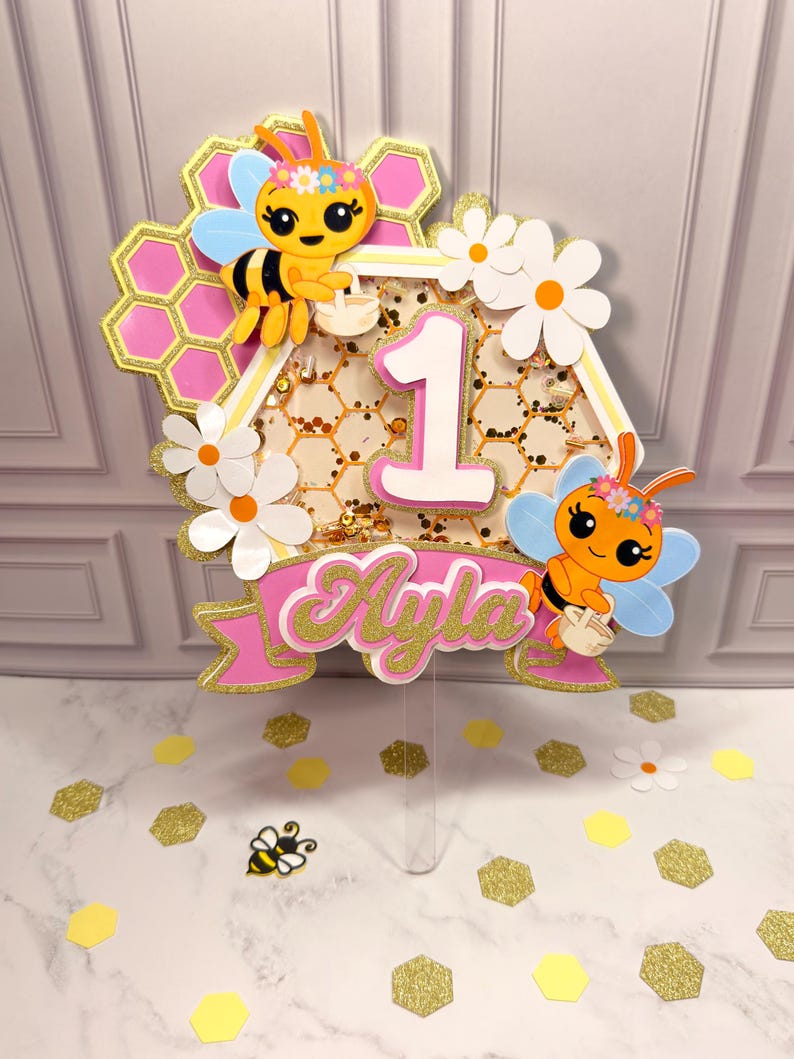 May include: A decorative cake topper featuring a hexagonal shaker filled with gold glitter, a large white number 1, and the name Ayla in pink script. The design includes two cartoon bees, pink honeycomb patterns, and white daisy flowers.