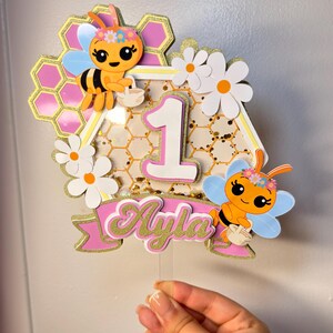 May include: A decorative cake topper featuring a central number one, two cartoon bees with flower crowns, white daisies, and a pink banner with the name Ayla in gold glitter. The design includes a honeycomb pattern and gold confetti accents.