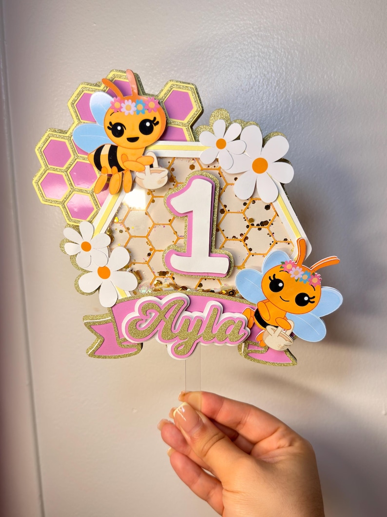 May include: A decorative cake topper featuring a central number one, two cartoon bees with flower crowns, white daisies, and a pink banner with the name Ayla in gold glitter. The design includes a honeycomb pattern and gold confetti accents.