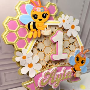 May include: A layered cake topper featuring a honeycomb background with pink accents, two cartoon bees with flower crowns, white daisies, and a central number one. A pink banner at the bottom displays the name Ayla in gold glitter script.