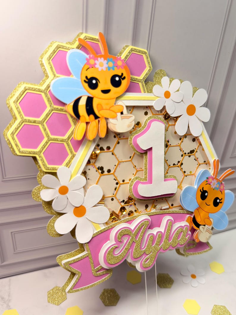 May include: A layered cake topper featuring a honeycomb background with pink accents, two cartoon bees with flower crowns, white daisies, and a central number one. A pink banner at the bottom displays the name Ayla in gold glitter script.