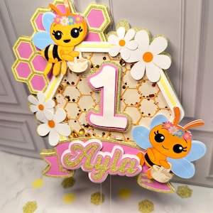 May include: A layered cake topper featuring a central hexagon with a honeycomb pattern and the number 1. It is decorated with two cartoon bees wearing flower crowns, white daisies, and a pink banner with the name Ayla in gold glitter script.