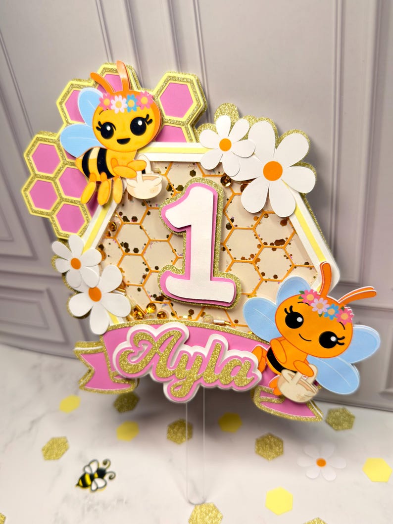 May include: A layered cake topper featuring a central hexagon with a honeycomb pattern and the number 1. It is decorated with two cartoon bees wearing flower crowns, white daisies, and a pink banner with the name Ayla in gold glitter script.
