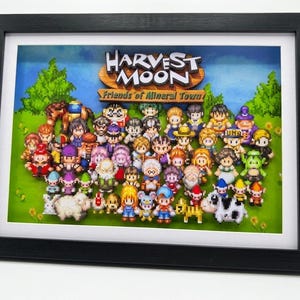 May include: A black framed shadow box featuring a multi-layered 3D paper art display of Harvest Moon: Friends of Mineral Town. The scene includes numerous pixelated character figures and animals on a green field with the game title at the top.