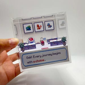 May include: A miniature diorama inside a clear acrylic box, featuring a pixelated scene with three framed images of cartoon characters. The scene includes a shop interior with a quote that reads, "Oak: Every journey begins with a choice."
