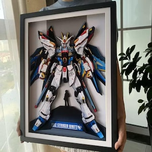 May include: Framed 3D paper art of a detailed robot, featuring white, blue, red, and yellow accents. The artwork includes a small figure and the text "AILE STRIKER UNIT" on a blue base. The robot is set against a gray background within a black frame.