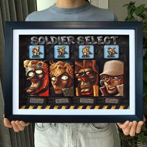 May include: Framed artwork featuring a "SOLDIER SELECT" screen from a video game. The artwork showcases four character portraits with names below: Marco, Eri, Tarma, and Fio. The frame is black with a white border.