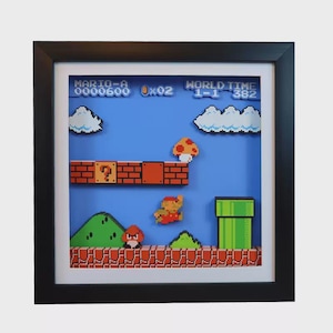 May include: Framed 3D artwork of a classic video game scene. The scene features pixelated characters, a blue sky, and brick platforms. The text "MARIO-A 0000600" and "WORLD TIME 1-1 382" are visible. The frame is black.