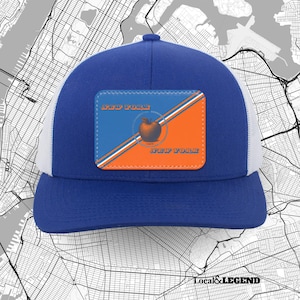 May include: A blue and white trucker hat featuring a rectangular patch on the front. The patch is divided diagonally into blue and orange sections with white stripes, displaying a central apple graphic and the text NEW YORK in orange lettering.