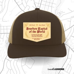 May include: A brown trucker hat with a tan mesh back and a rectangular patch on the front. The patch is cream-colored with dark red text reading Bourbon Capital of the World, 100 Proof, Sour Mash, and Kentucky Straight Bourbon Whiskey.