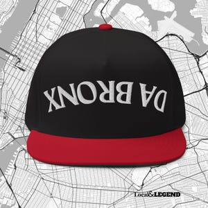 Puede incluir: Una gorra de béisbol negra con visera plana roja y un botón rojo en la parte superior. La parte frontal de la gorra presenta el texto DA BRONX bordado en hilo blanco, colocado al revés en el centro de la corona.