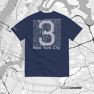 Puede incluir: Una camiseta azul marino de manga corta con un gran número 3 blanco superpuesto sobre un mapa de Nueva York. El texto The House That Ruth Built aparece sobre el número, con New York City debajo. La palabra Legend está en la manga.