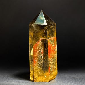 Op de afbeelding: Een facetgeslepen, goudgeel kristal met een puntige top. Het kristal heeft een rood-oranje insluitsel dat verticaal door het midden loopt. Het kristal staat tegen een donkere achtergrond.