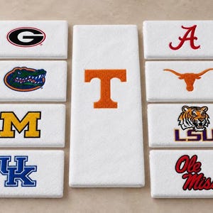 May include: A collection of white rectangular towels featuring various embroidered collegiate sports logos, including the Georgia G, Florida Gator, Michigan M, Kentucky UK, Alabama A, Texas Longhorn, LSU tiger, Ole Miss text, and a large orange T.