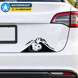 以下が含まれることがあります： 白い車両の後部に貼られた黒いビニール製のデカール。このデカールは、丸い頭と垂れた耳を持つ漫画の犬のグラフィックイラストで、暗い三角形の形の上から顔を出し、片手を振っている様子が描かれています。