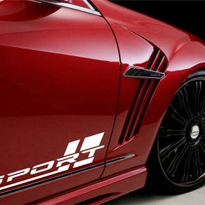 Op de afbeelding: Een glanzend rood zijpaneel van een auto met een witte vinyl sticker met het woord SPORT in dikke hoofdletters, vergezeld van twee rechthoekige vormen. Het voertuig is voorzien van een zwart meerspaaks wiel en een zwarte zijventilatie-afwerking.
