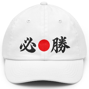 May include: A white baseball cap featuring a black brushstroke-style Japanese kanji character on the left, a solid red circle in the centre, and a black brushstroke-style Japanese kanji character on the right, with a curved brim and eyelets.