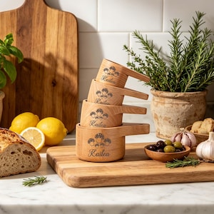 Può includere: Una pila di quattro misurini in legno con manici, ognuno inciso con il nome "Kailea" e un disegno di funghi. Le tazze sono su un tagliere di legno, con limoni, pane ed erbe aromatiche sullo sfondo.