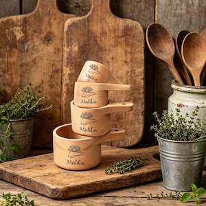 Könnte beinhalten: Ein Stapel von vier Holzmessbechern mit Griffen, jeweils mit dem Namen "Malika" und einem Blumendesign graviert. Die Becher stehen auf einem Holzschneidebrett, mit Kräutern und Holzlöffeln im Hintergrund.