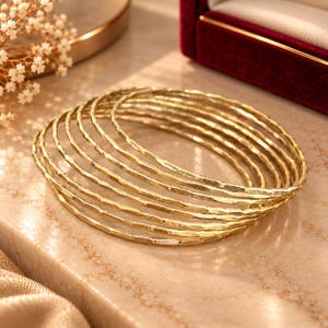 May include: A set of seven gold-toned bangles featuring a hammered, textured surface. Each circular bracelet has a thin, uniform width and a reflective metallic finish, creating a stacked appearance when arranged together in a circular formation.